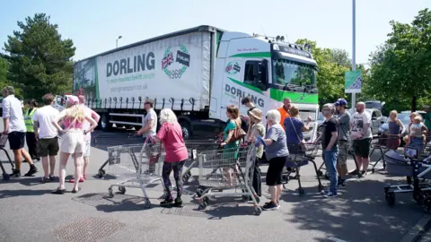 PA Media People are lining up to get bottled water at a car park. Some are pushing trolleys and a lorry is in the background.