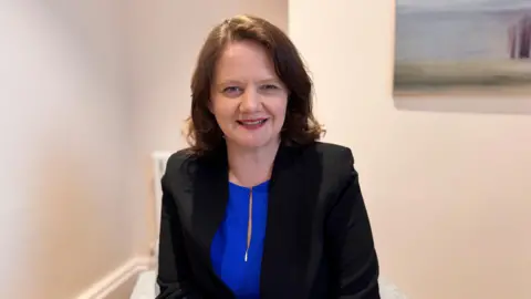 Ellen O’Donoghue who is looking at the camera and smiling. She has a blue top with a zip and a black jacket. Her hair is brown. 