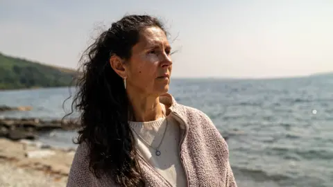 Catherine - with long dark hair standing on a rocky shoreline beside a large body of water, wearing a light-coloured fleece jacket and looking toward the distance. The coastline and hills are visible in the background under a bright, clear sky.