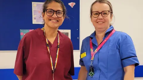 NHS Humber Health Partnership Image of two female health professionals standing inside a hospital, One has tied back dark hair and glasses and is wearing a red top . The second woman has fair hair and is wearing a blue top. Both have lanyards.