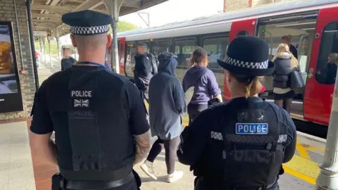 The picture has been taken on a train station and there are several people getting on the train. There are two police officers, a male and female, who have their backs against the camera.
