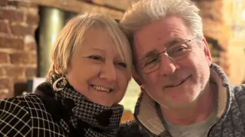 Cambridgeshire Police Gordon Fox sitting beside a woman with short fair hair. He is on the right and is wearing a grey zipped fleece with the collar raised. He has white hair and is wearing metal framed glasses. His head is touching that of the woman's and they are both smiling. 