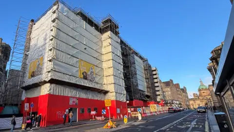 BBC A white plastic sheet covering scaffolding around a hotel building Edinburgh. The sky above is blue. The lower half of the scaffold is covered by red, wooden panelling.