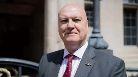 Ian Cooper, a man wearing a dark grey suit and a dark red tie, is standing outside Stafford County Buildings. He is also wearing a white shirt and a pin badge on his lapel.
