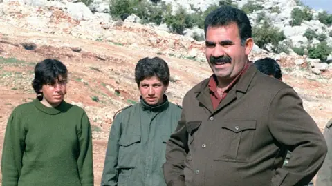 Getty Images stands ocalan to the camera with his hands on the hips. Male fighters behind him.