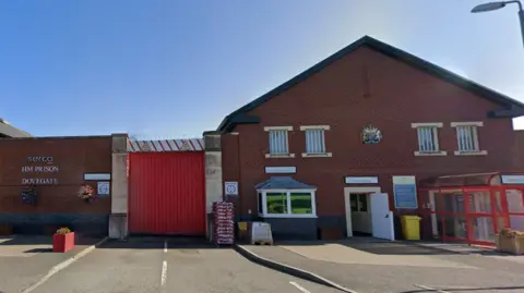 Google The outside of a red-brick building with large red gate. It has Serco HM Prison Dovegate in letters on a wall on the left of the gate and an official crest in between second floor windows to the right. Crates containing bags are piled up by the gate and there are open entrances to the building on the right of the gate. The sky is blue.