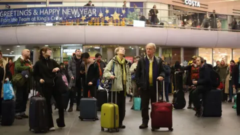 PA Media Rail passengers at King's Cross station in London wait for their trains wearing winter coats and carrying their luggage. A sign reading "Greetings and Happy New Year from the team at King's Cross" is hanging on a gallery above the station concourse. 