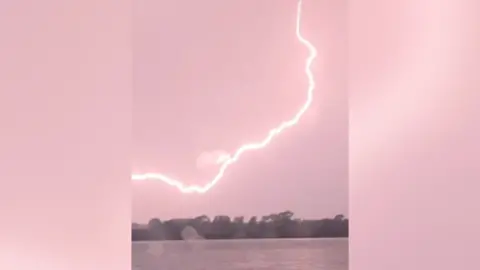 Image of a large arc of lightning streaking across the sky. The bolt is bright enough to tinge the sky a reddish pink. 