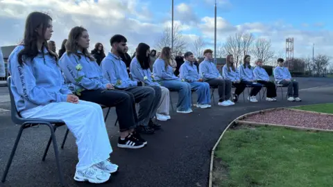 The pupils are wearing light coloured tops and are sitting on chair outside the school.