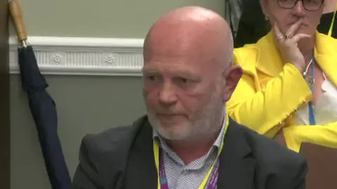 Dr Chris Hagan sits in a meeting, he is bald with grey facial hair. He is wearing a black blazer, patterned shirt and has a purple and yellow lanyard around his neck.