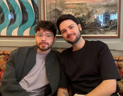 Joe Diaz Two men with brown hair one wearing a purple t shirt and another in a black t shirt sitting in a restaurant smiling.
