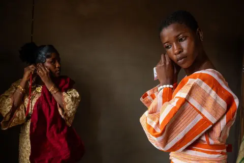 Toyin Addokun / AFP / Getty Images Two women put on earrings as they get ready. One is in an orange and white stripped outfit the other is in a cream and maroon dress - Friday 31 October 2025.