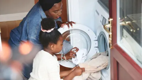 A mother and daughter put laundry into a washing machine together in their home