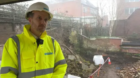 Paul Maddison, from Ramboll, is photographed in front of Chester's walls. He is wearing a white hard hat and a yellow hi-vis jacket bearing his company's logo.