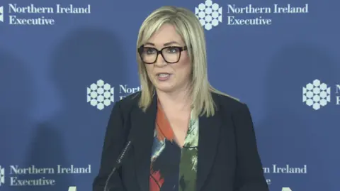 The first minister wearing a dark suit and multicolour shirt. Behind her is a blue background with the Northern Ireland executive logo.