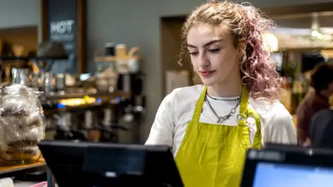 Getty Images A young woman working on a till in a cafe wearing a yellow apron