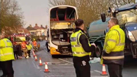 Picture of the crash scene with the double decker bus with smashed windows and a blue tractor with tanker. Emergency services stand around and a fire engine in the background.