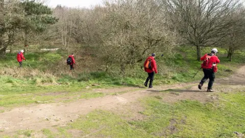 Four volunteer rescuers in red tops and black trousers walk through a woodland area.