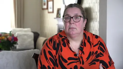 Barbara Cunningham, 53, sitting in a dining chair wearing glasses and a red and black blouse, looks into the camera with a downcast expression. 