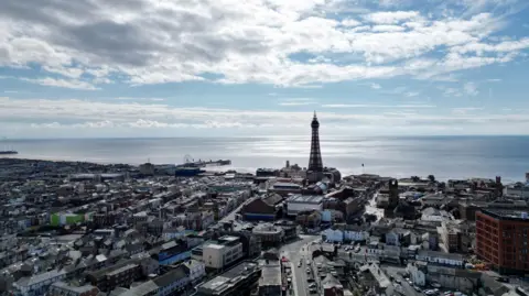 An overhead image of the seaside town of Blackpool, with the Tower on the horizon, buildings in the front of the image and the sea in the background.