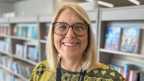 Aimee Dexter/BBC Kerry Murray is standing in front of a grey book shelf which has several books on it. She has shoulder length blonde hair and is wearing a yellow and black T-shirt and a black lanyard. 