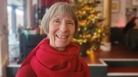 Alexandra Pickford is sitting in a cafe dressed in a red top and red scarf. She has short, grey hair and is smiling. There is a Christmas tree lit up behind her.