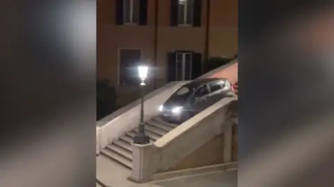 A grey car drives down the Spanish Steps at night with a street lamp illuminated next to it.