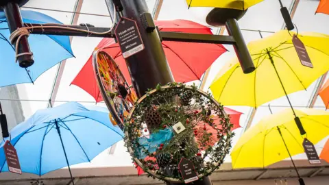 Charter Walk Shopping Centre blue red and yellow umbrellas hang on the ceiling of the shopping centre and a bike covered in foliage, including a pine cone, is attached to a post