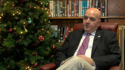 Jonathan Le Tocq, vêtu d'une veste de costume sombre, d'une chemise blanche et d'une cravate rose, assis les jambes croisées dans un fauteuil en cuir rouge à côté d'un sapin de Noël décoré