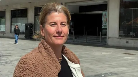 Sian Ridpath is wearing a black top, a cream cardigan and a brown fleece jacket and is standing in Armada Way