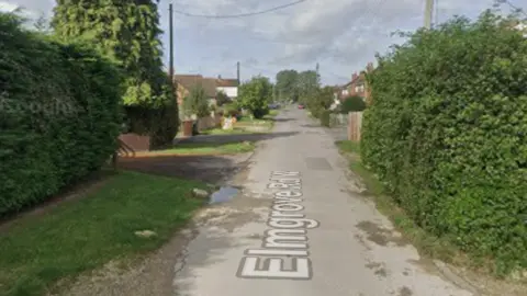 A street view of a country road with houses and hedges on either side.