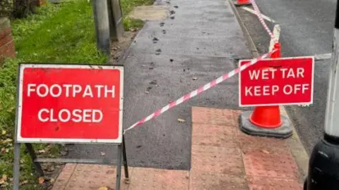 The two signs read 'Footpath Closed' and 'Wet Tar Keep off'. However, behind the signs are footprints in the tar.