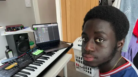 A 20 year old student sits next to a keyboard, speaker and a laptop in his university room