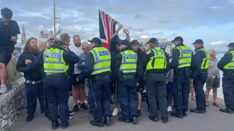 Seven riot police wearing black jackets and black trousers with bright yellow police vests holding a group of men back who are waving a union jack flag of a white pole. 