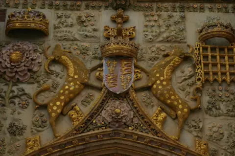 Christopher Hilton/Geograph An embossed coat of arms over a stone gateway. It features a shield with a crown on top of it, and two yales standing on their hind legs "supporting" the shield. On the left of a yale is a crown above a flower. On the right of the other yales is a crown above a portcullis.