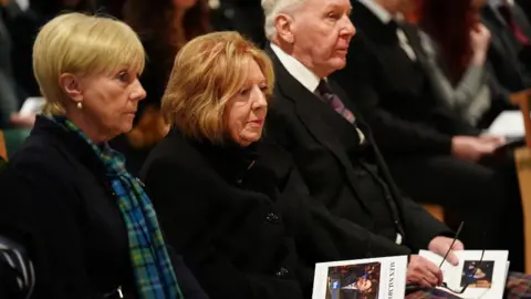 PA Media Moira Salmond, with ginger hair and dressed in black, sits in the front row of a memorial service for her late husband 