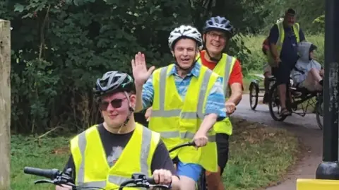 Bike riders smile at the camera as they cycle on a path. They wear helmets and hi-vis jackets as they cycle.