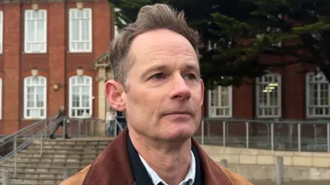 George Carden/BBC A man with brown hair wearing a brown coat. He is standing in front of a red brick building.