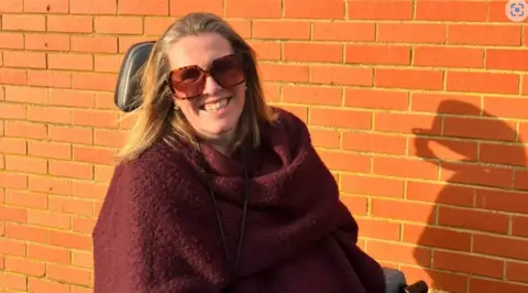 Handout Woman in a wheelchair with a purple shawl and sunglasses. She is in front of a brick wall smiling widely