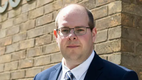 Conservative Party A picture of Conservative Hertfordshire Police and Crime Commissioner Jonathan Ash-Edwards wearing a blue suit jacket with a blue and white striped tie.