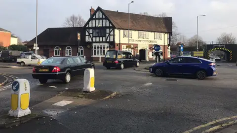 A roundabout showing a hearse travelling straight over the island, followed by another black car. 