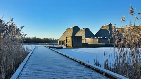 Ilse's Imagery A wintry scene of Brockholes nature reserve. A wooden walkway dusted in snow leads to a modern wooden structure visitor centre with pitched roofs located on a pontoon in a frozen lake.