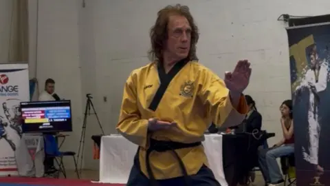 Mike Pejic Mike Pejic has long brown hair and is wearing a yellow martial arts top and black trousers. He is standing side-on in a taekwondo stance with his left arm raised up and right arm by his stomach.