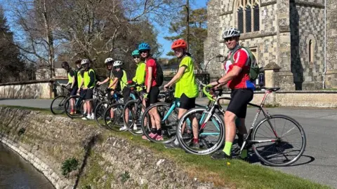 Mark Ball A group of teenagers and their teacher, Mark Ball. They are standing in a line with bicycles. They are all wearing biking gear and looking at the camera.