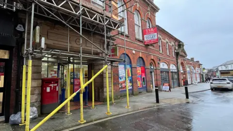 ELLEN KNIGHT/BBC Photo of the exterior of the Post Office. It's in a large red brick building, and has two red post boxes outside the front doors. A red and white sign hangs over the entrance, reading 'Post Office.' Much of the entrance is slightly obscured by scaffolding, which is metal and has yellow material wrapped around it. The pavement is wet, suggesting recent rain, and the sky is grey and overcast.