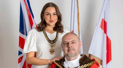 A bald man in mayoral robes sits in front of a Union Jack flag and several England flags. Stood behind him with her hands on his shoulders is a young woman with brown shoulder-length hair in a white dress, with a mayoress's chain around her neck.
