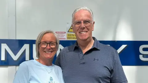 Lorna and Roger standing side by side. Lorna has short blonde hair, black rounded glasses and is smiling at the camera as she wears a blue t-shirt. Roger has short grey hair, blue striped shirt and silver metal glasses.
