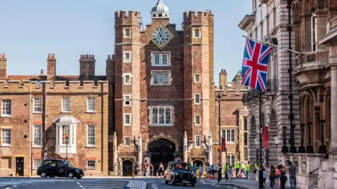 The exterior of St James's Palace,  a Tudor brick building, on a sunny day. There are black taxis on the road in front of the palace and people milling about around the entrance.  