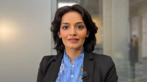 Dr Nita Mandhar, the medical director of NEMS. She is a young, South Asian woman with short black hair. She is wearing a black blazer and with a blue floral blouse.