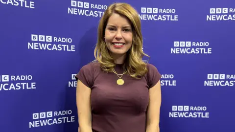 Kim McGuinness smiling into the camera in front of a purple BBC Radio Newcastle backdrop. She has curled, blonde hair which falls just below her shoulders and is wearing a large gold necklace and a purple t-shirt. 